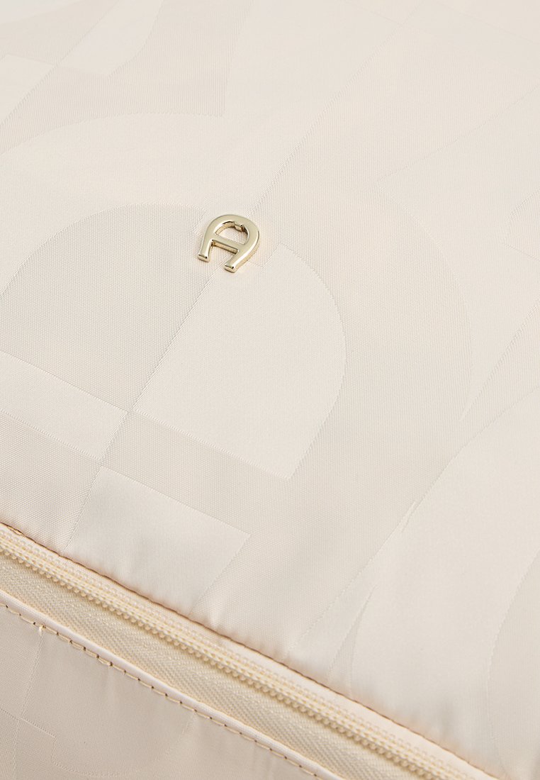 Beige Stofftasche mit einem dezenten Muster aus überlappenden Formen, ausgestattet mit einem goldenen Metall-Logo und einem cremefarbenen Reißverschlussdetail entlang der Kante.