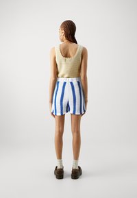 Top de tirantes de punto de ganchillo beige con bordes ondulados, combinado con pantalones cortos blancos con rayas horizontales azules. Zapatos de plataforma marrones complementan el conjunto.