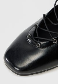 Scarpa piatta in pelle nera con punta quadrata e lacci elastici incrociati passati attraverso occhielli in metallo su uno sfondo bianco.