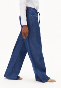 Weite Jeanshose aus dunkelblauem Denim, mit hohem Bund, vorderen Taschen und Kordelzugverschluss. Glatte Textur, traditionelle Nähte.