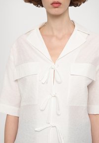 Witte kortekmouwenshirt met een gestructureerde stof, knoopsluiting met strikjes en twee flapzakken op de borst.