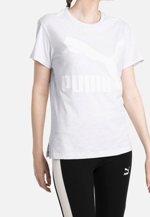 Personne portant un t-shirt blanc Puma et des leggings noirs Puma avec des bandes blanches sur les côtés, debout devant un fond uni.