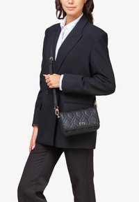 Bolso negro acolchado con una correa desmontable, que presenta herrajes dorados y un detalle del logo. La superficie de cuero tiene un acabado texturizado.