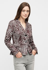 Νεαρή γυναίκα με μακριά καστανά μαλλιά, ντυμένη με σακάκι με σχέδιο λεοπάρδαλης και ανοιχτό γκρι τζιν, στέκεται με τα χέρια στις τσέπες μπροστά σε ένα απλό φόντο.