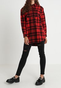 Camicia a scacchi rossa e nera in flanella con maniche lunghe e due tasche sul petto, indossata su jeans skinny neri strappati e mocassini neri.