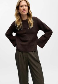 Donkerbruin gestructureerd breisweater met brede mouwen, gecombineerd met losse groene broeken. De sweater heeft een geometrisch patroon.