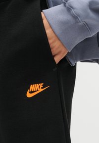 Χέρι στην τσέπη μαύρου παντελονιού Nike με φωτεινό πορτοκαλί λογότυπο, φορώντας μπλε φούτερ με εμφανές μανσέτα και λεπτομέρειες φερμουάρ.