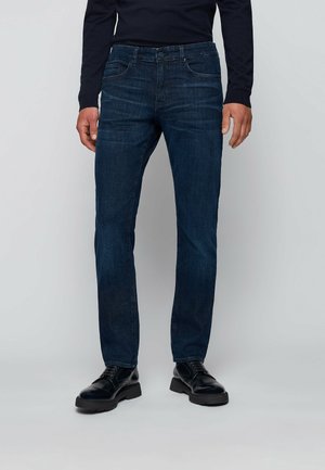 Man draagt donkerblauwe slim-fit jeans, zwarte leren veterschoenen en een zwart hemd met lange mouwen tegen een effen achtergrond.