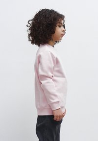 Felpa rosa chiaro con polsini a costine e colletto tondo, abbinata a jeans scuri. Il bambino è in profilo su uno sfondo neutro.