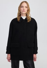 Cappotto di lana nera con colletto, chiusura frontale a bottoni e due grandi tasche a patta. Il design è morbido e presenta dettagli sulle maniche.