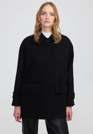 Czarny wełniany płaszcz z kołnierzykiem, z przodu zapinany na guziki i dwoma dużymi kieszeniami na klapki. Krój jest luźny z akcentami na rękawach.