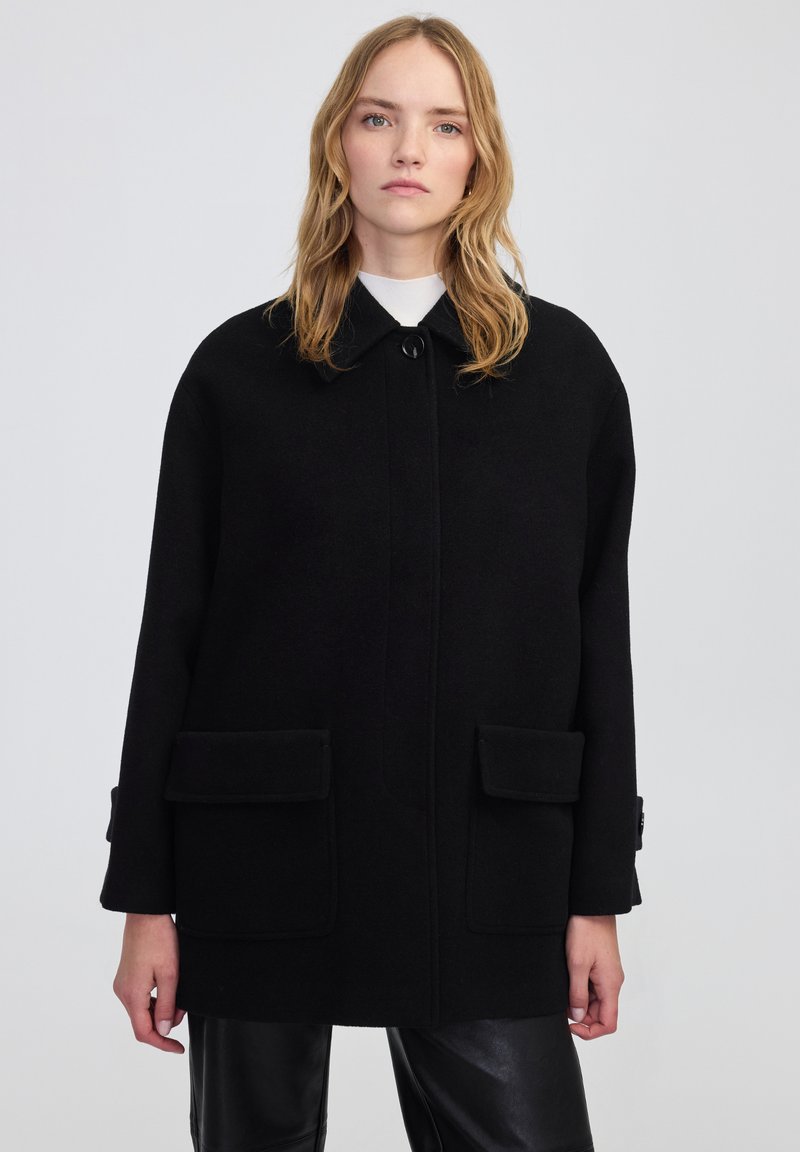 Cappotto di lana nera con colletto, chiusura frontale a bottoni e due grandi tasche a patta. Il design è morbido e presenta dettagli sulle maniche.