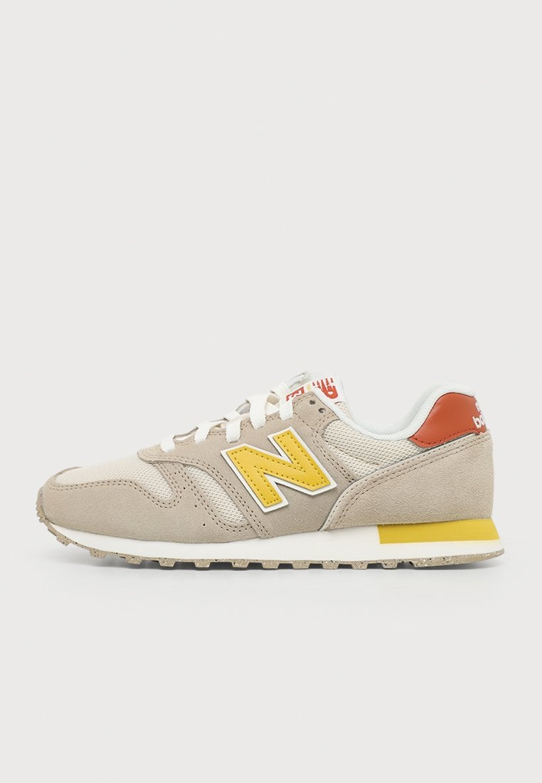 Zapatilla deportiva beige de New Balance con cordones blancos, un logotipo amarillo en el lateral y un detalle amarillo en el talón, mostrada de perfil sobre un fondo sencillo.