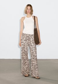 Witte tanktop, beige broek met een donkerbruin spikkelpatroon, bruine schoudertas, platte sandalen, staand op een grijze vloer tegen een lichte achtergrond.