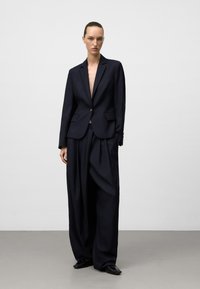 Blazer aderente blu navy con reverse a punte, chiusura a un bottone e tasche a pattina, abbinato a pantaloni larghi a vita alta.
