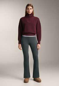 Burgundské pletené svetr s vysokým límcem a texturovaným vzorem, spárovaný s tmavozelenými jemně rýhovanými kalhotami s rozšířenými nohavicemi a tanovými slip-on botami.