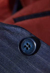 Marinblått tyg med slät textur, som har en rund knapp med fyra hål. Bakgrunden består av ett rödbrunt tyg.