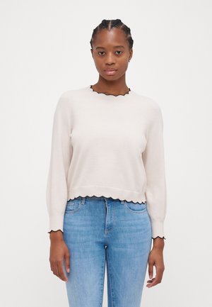 Pull en tricot beige clair avec un col et un ourlet noir à volants, à manches longues, porté sur un jean bleu clair. Texture douce et coupe décontractée.
