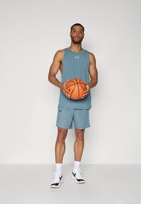 Atleta vestindo uma regata azul claro e calções, segurando uma bola de basquete laranja. Ele está em pé sobre um fundo simples, usando ténis brancos e pretos.