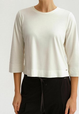Weißes, kurzes Langarmoberteil aus glattem Material mit rundem Ausschnitt und Nähten. Getragen mit schwarzen Bauchtaille-Hosen mit einem Bindeband.