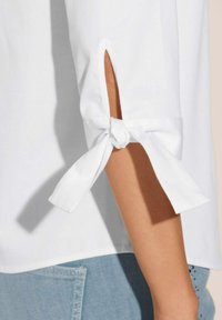 Witte blouse met een mouw met uitsnijding en een strikdetail. Zachte stof, losse fit, geaccentueerd door een lichte, casual denim achtergrond.