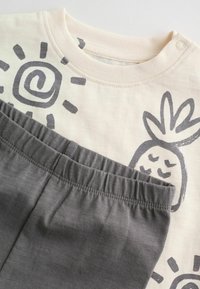 Svetlo bež majica sive ilustracije ananasa in vzorcev sonca, združena s temno sivimi hlačami z elastičnim pasom, mehka bombažna tekstura.