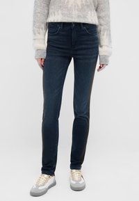 Sötétkék farmernadrág, slim fit szabással, fekete oldalsó panelekkel és klasszikus zsebekkel. Krém- és szürkeszínű sportcipővel párosítva.