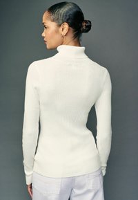 Weißer gestreifter Rollkragensweater mit langen Ärmeln, der eine figurbetonte Silhouette und eine strukturierte Oberfläche aufweist, auf einem Modell von hinten präsentiert.