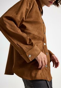 Camicia in suede marrone con vestibilità rilassata, chiusura a bottoni a pressione e maniche lunghe. Presenta un orlo curvo e dettagli in metallo argentato.
