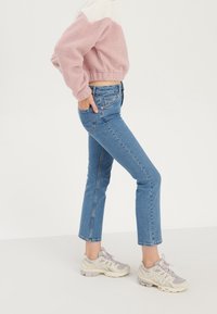 Hellrosa-cremefarbener Fleecepullover mit elastischem Saum, kombiniert mit eng anliegenden blauen Jeans und beigen Sneakers mit grauen Akzenten.