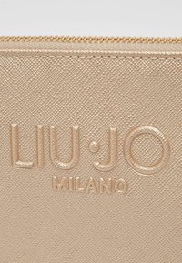 Pochette en simili cuir doré métallisé avec surface texturée. Présente un logo embossé "LIU·JO MILANO" et une fermeture éclair dorée sur le dessus.