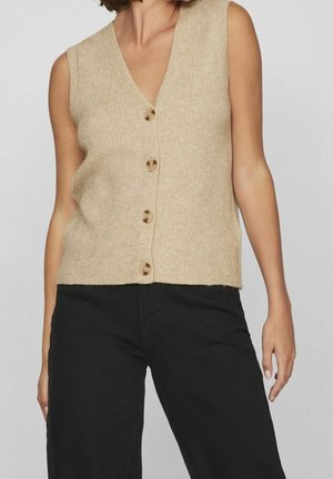 Gilet sans manches en maille beige avec quatre boutons, porté avec un pantalon noir, montrant le torse et les bras sur un fond uni.