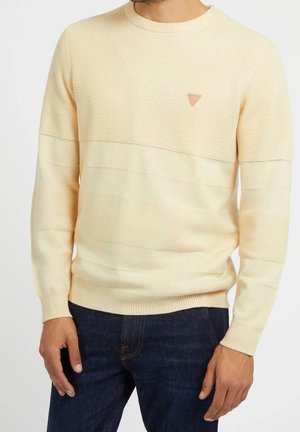 Beiger langärmeliger Strickpullover mit strukturierter oberer Hälfte und dreieckigem Logo auf der linken Brust, getragen mit dunkelblauen Jeans.