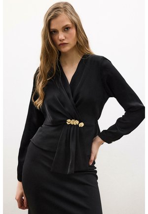 Femme portant une robe portefeuille noire à manches longues avec des ornements floraux dorés à la taille, posant avec une main sur la hanche devant un fond uni.