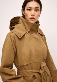 Veste imperméable marron avec un col haut, des boutons-pression, une taille ceinturée et des poches latérales. Le tissu est lisse avec une finition mate.