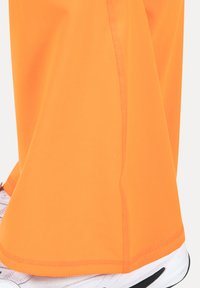 Oranssit urheiluhousut, joissa on sileä pinta, suoraleikkaus ja kaksinkertaiset saumat, yhdistettynä valkoisiin urheilukenkiin.