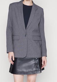 Blazer gris a medida con solapas con muesca, cierre de un solo botón y dos bolsillos laterales; combinado con una minifalda de cuero negro con botones.