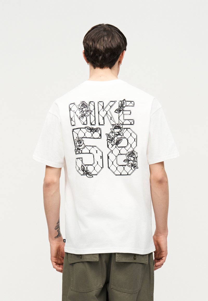Άτομο που φοράει λευκό t-shirt με το «NIKE 58» περίγραμμα σε μαύρο λουλουδάτο και αλυσίδα σχέδιο, συνδυασμένο με παντελόνι ελιάς.