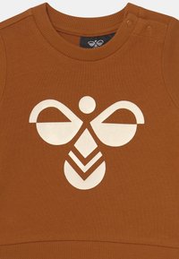 Brun ribbad sweatshirt med en krämfärgad abstrakt design som har cirklar och geometriska former. Inkluderar tryckknappar på axeln.
