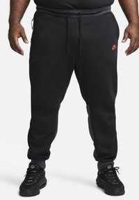 Schwarze Nike-Sweatpants mit slim fit, elastischem Bund und rotem Logo. Besitzt eine glatte Textur und seitliche Taschen. Kombiniert mit schwarzen Sneakers.