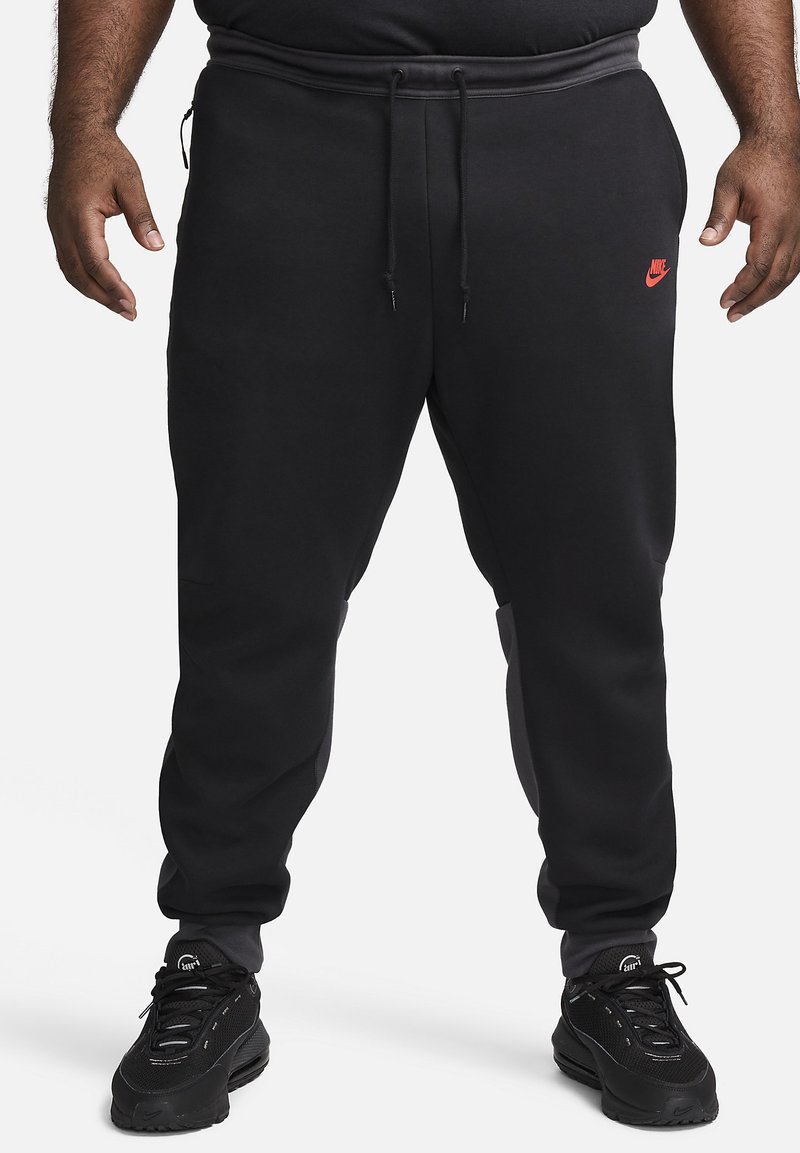 Schwarze Nike-Sweatpants mit slim fit, elastischem Bund und rotem Logo. Besitzt eine glatte Textur und seitliche Taschen. Kombiniert mit schwarzen Sneakers.
