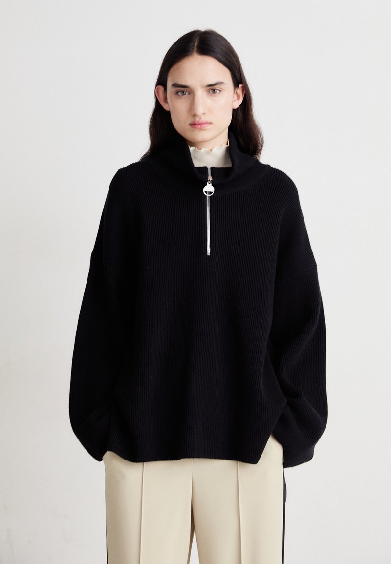 Schwarzer gerippter Strickpullover mit hohem, umgeschlagenem Kragen und silbernem Reißverschluss. Hat übergroße Ärmel und einen lockeren Schnitt. Kombiniert mit cremefarbenen Hosen.