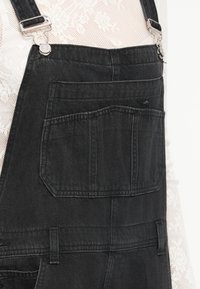 Črne denim kombinezoni imajo nastavljive naramnice, sprednji žep in rahlo teksturo. Material izgleda trpežno z mat površino.