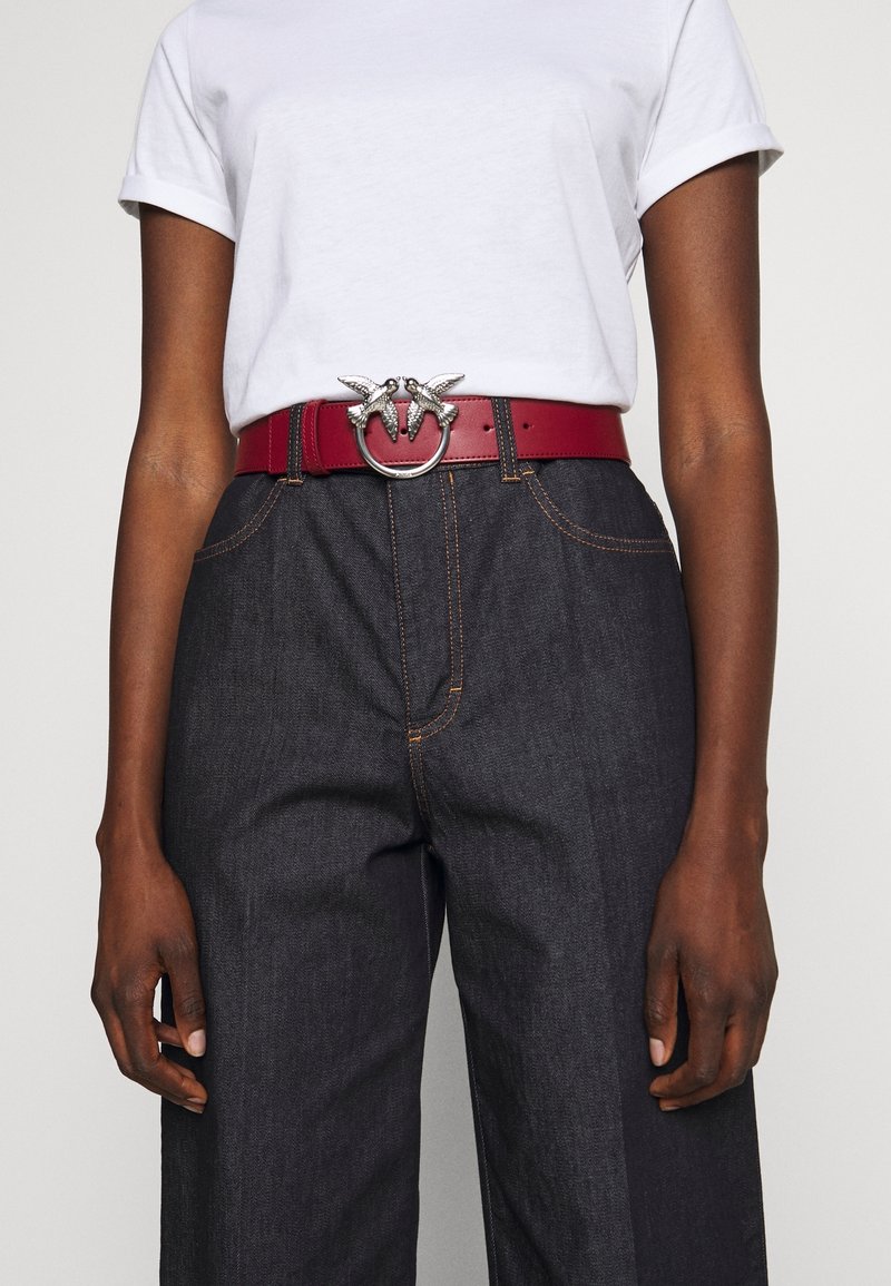 Vue à hauteur de taille d’une personne portant une chemise blanche à manches retroussées, un jean sombre taille haute et une ceinture rouge avec une boucle argentée en forme d’oiseau.