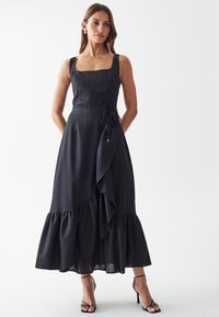 Black linen dress with square neckline, side tie detail, and layered ruffle hem. Worn with strappy black heels.