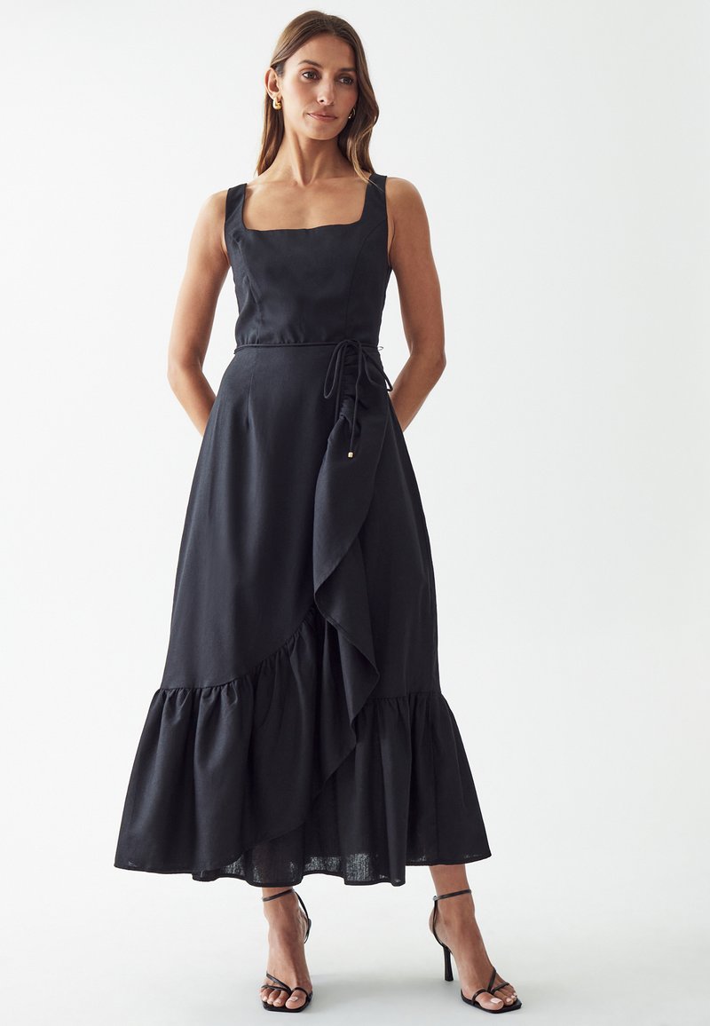 Black linen dress with square neckline, side tie detail, and layered ruffle hem. Worn with strappy black heels.
