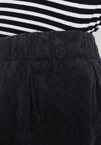 Pantalones de pana negros con un patrón texturizado, que presentan una cinturilla ancha y elástica y pliegues bien definidos para mayor volumen.