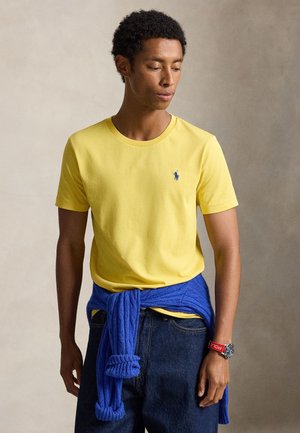 Homme portant une chemise jaune à manches courtes, un pull bleu noué à la taille, un jean foncé et une montre rouge, regardant vers le bas à sa gauche.