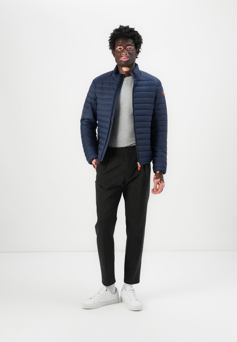 Giacca trapuntata blu navy con colletto alto, maglione grigio, chinos neri e sneakers bianche. Presenta un tocco di arancione sulla cerniera.