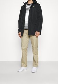 Czarna wodoodporna kurtka z kapturem, zapinana na guziki z przodu, z dwoma bocznymi kieszeniami, w połączeniu z koszulą w paski i beżowymi spodniami, dopełnia stylizację.